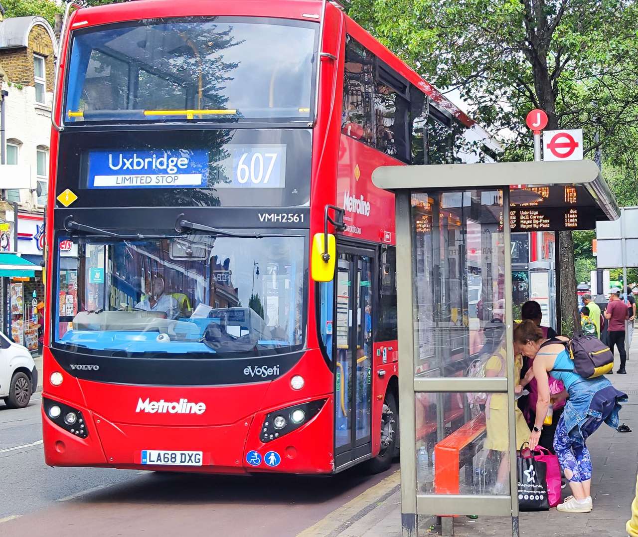 A red double-decker bus showing Uxbridge pulling into a bus shelter on a tree-lined street
